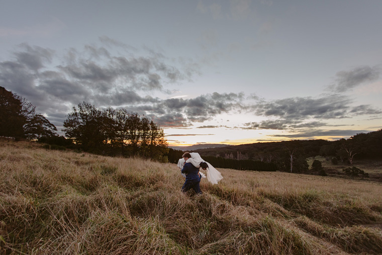 Groom carrys bride through a long grass field.